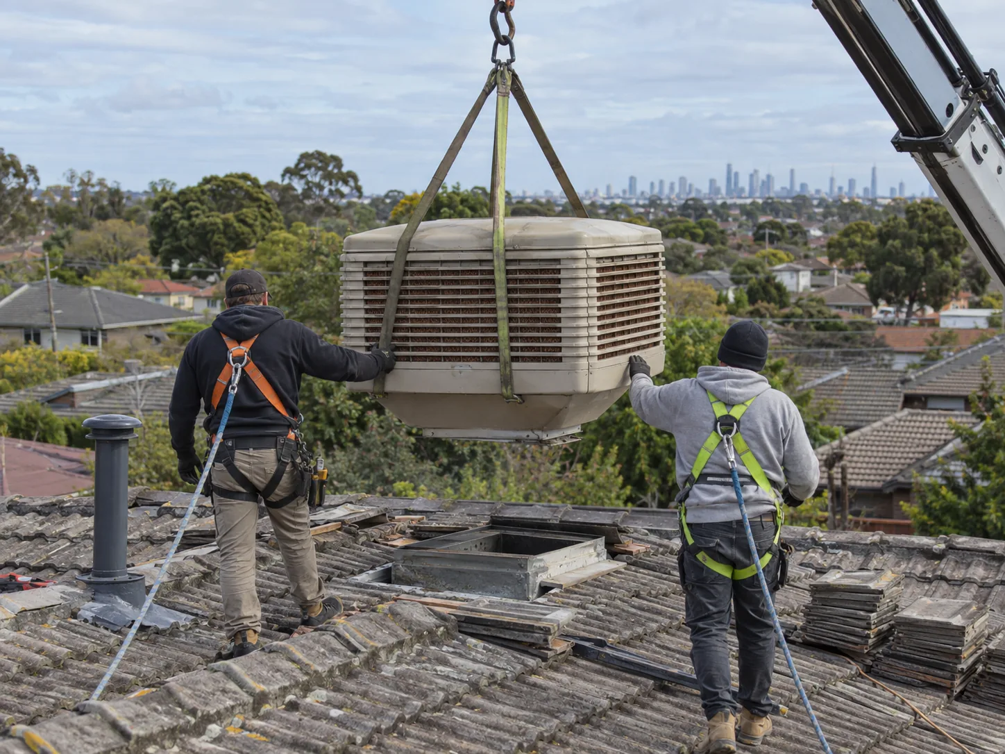 An evaporative cooler being craned off a tiled roof, two technicians guiding it