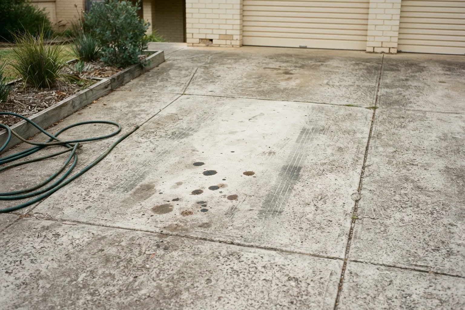 Concrete driveway with dark oil drip stains and tyre marks, a garden hose coiled at the side